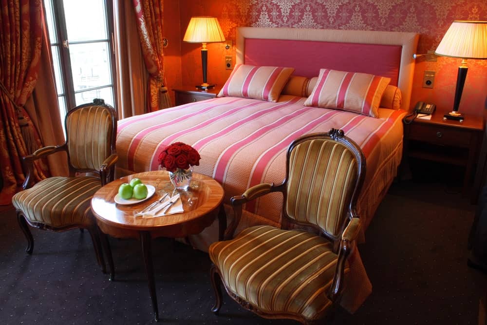传统床的粉色床单和粉色软垫床头板与粉红色图案的墙纸融合在一起。两盏台灯投射出温暖的黄色灯光，从窗户射进来的自然光增强了这一效果。