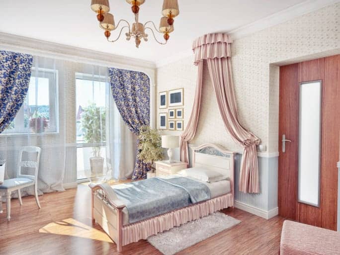 这是一个优雅的维多利亚风格的可爱的女孩卧室。木制床头板上方有一个装饰性的粉红色窗帘，与床架的侧裙相匹配。这与有图案的壁纸和硬木地板搭配在一起。
