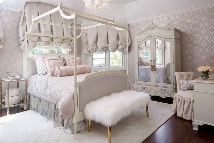 这间维多利亚风格的卧室里，迷人的白色木制四柱床以床单、壁纸和窗帘的浅灰色元素为主。床脚的长凳上有一个白色的毛茸茸的靠垫和金色的腿，在白色图案的地毯的衬托下显得很突出。