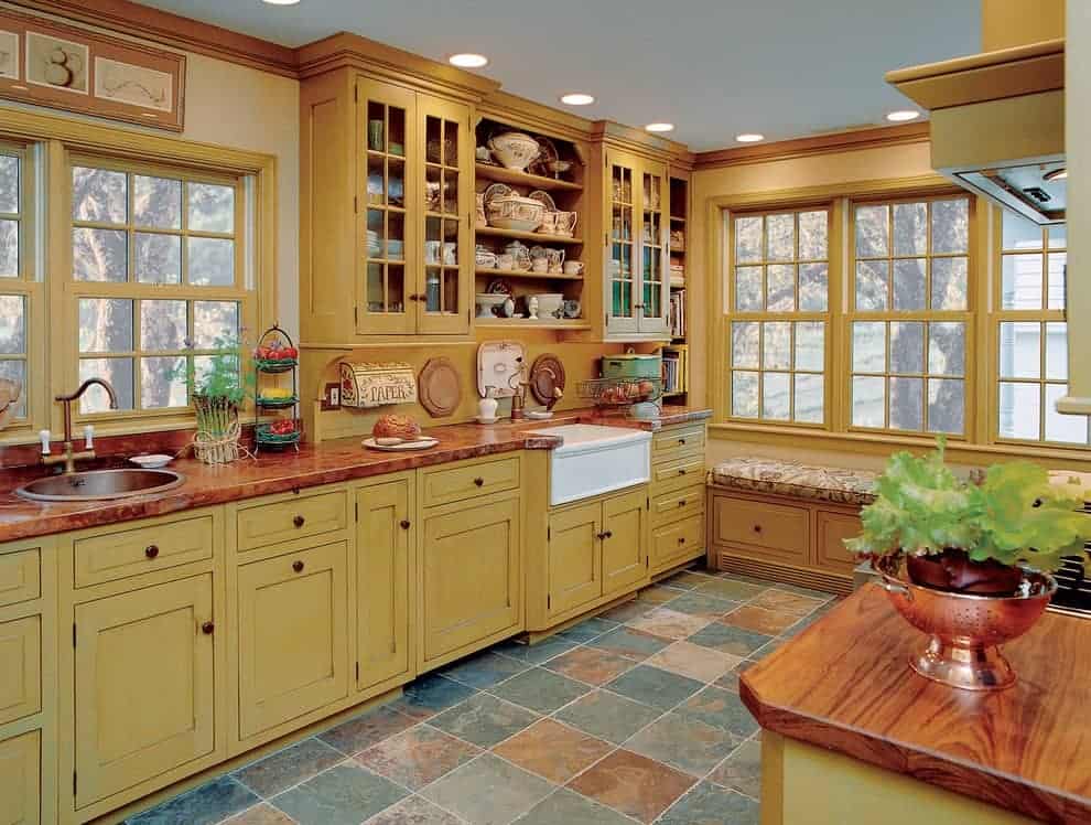农舍厨房与石灰石地板和框窗俯瞰户外风景。它包括黄色和玻璃的前柜，以及一个顶部有花卉靠垫的座位角落。