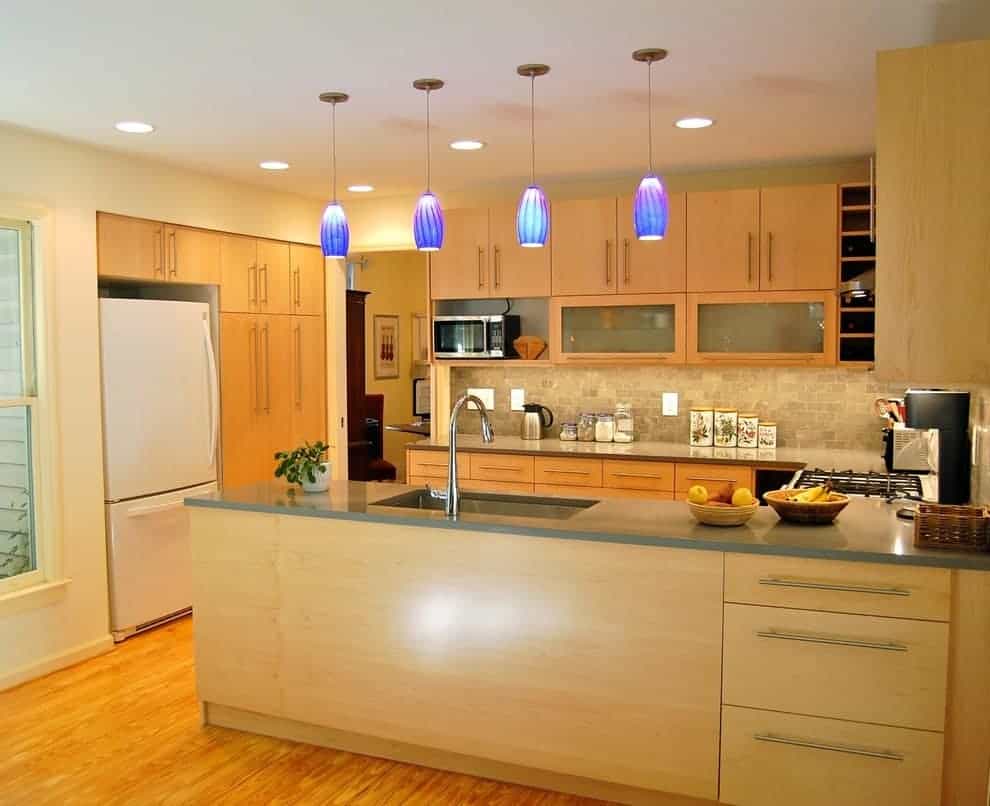 现代厨房，白色冰箱和轻木橱柜与半岛相辅相承，半岛配有下置水槽和鹅颈水龙头。它由蓝色吊坠和凹形天花板灯照亮。