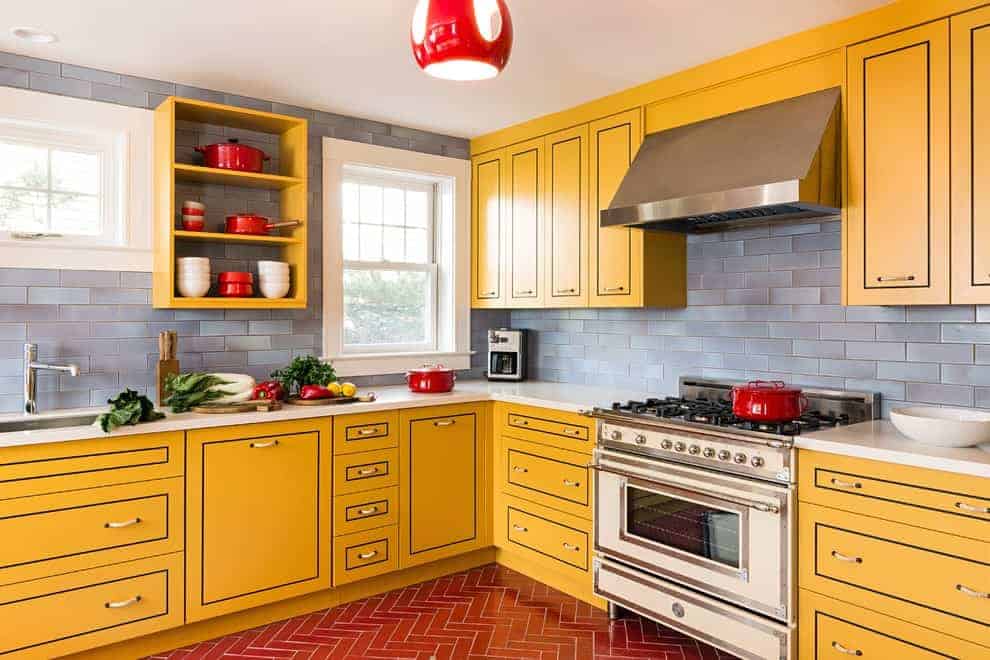 红色的照明灯具照亮了这个厨房，灰色的地铁瓷砖后挡板和黄色的橱柜由黑色的装饰突出。它有白色框窗和人字形瓷砖地板。