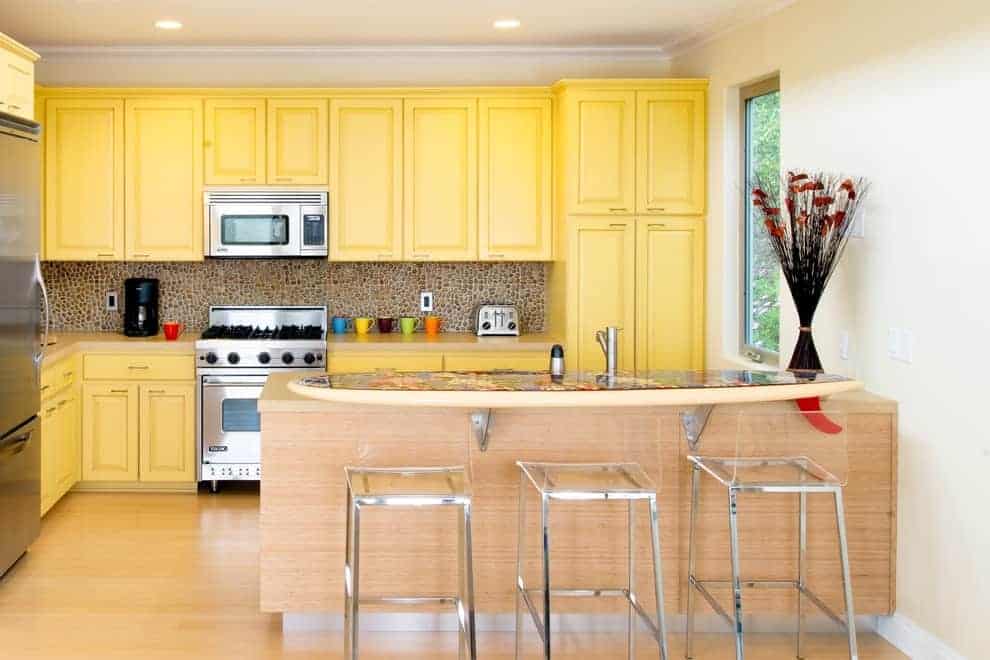 这个厨房展示了不锈钢电器和黄色的橱柜，并配有鹅卵石后挡板。它包括一个浅色的木质半岛，带有一个凸起的就餐柜台，上面排列着玻璃酒吧椅。