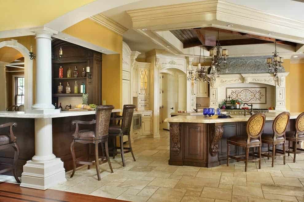 豪华厨房提供一个酒吧和花岗岩顶部岛，在石灰岩地板上排列着圆形靠背椅。它包括一间华丽的烹饪凹室和悬挂在托盘天花板上的复古枝形吊灯。
