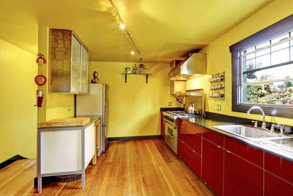 红色橱柜与厨房的黄色墙壁形成了鲜明的对比，厨房里有黑色的台面和不锈钢用具。它由轨道灯和铝框窗的自然光照亮。