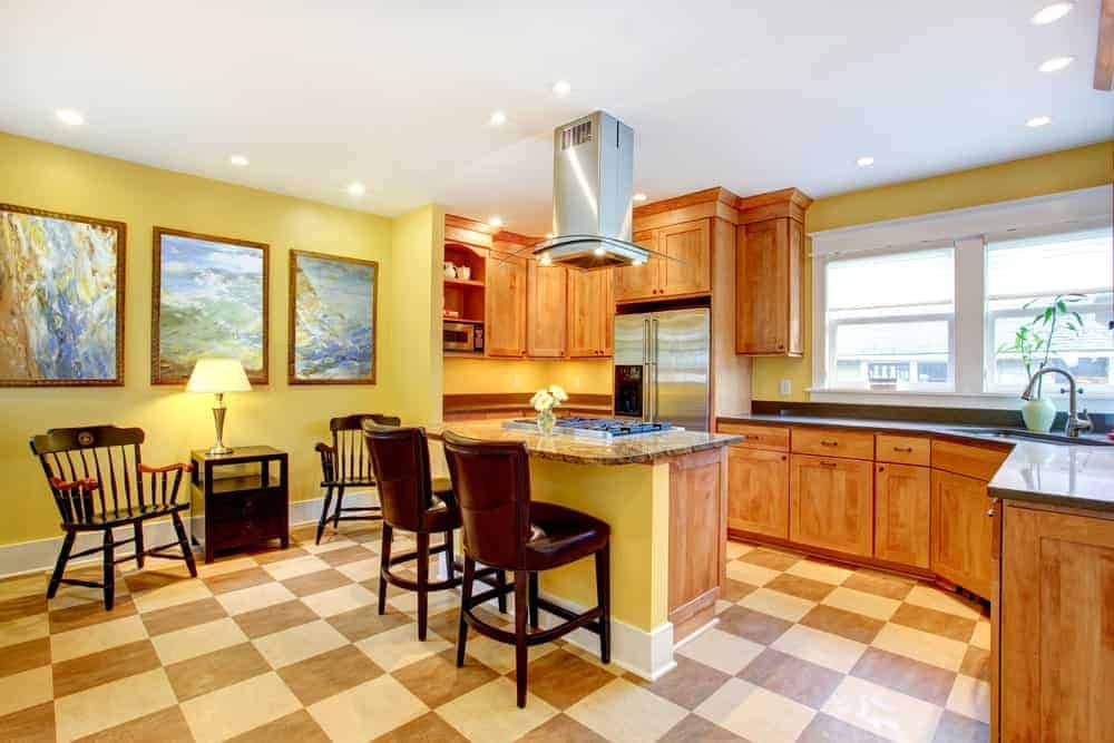 明亮的厨房设有木制橱柜和一个装有内置灶台的小厨房岛。旁边有一个座位区，黄色墙上的抽象画突出了它。