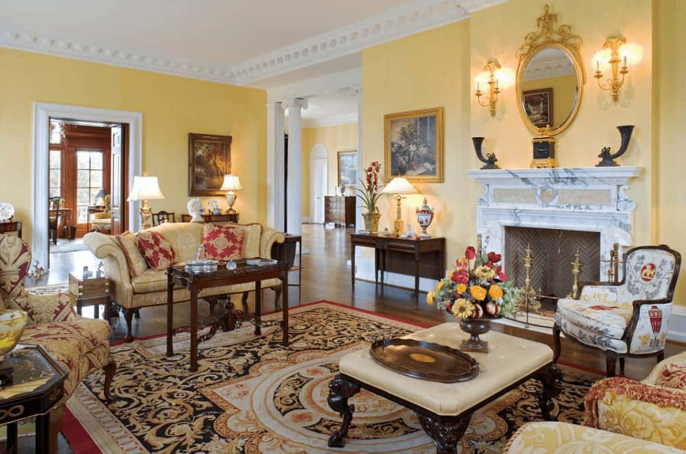优雅的客厅设有优雅的座位和一个雪佛龙壁炉框架与白色大理石壁炉架。它包括一个经典的区域地毯和华丽的镜子安装在黄色的墙上衬着白色的皇冠造型。