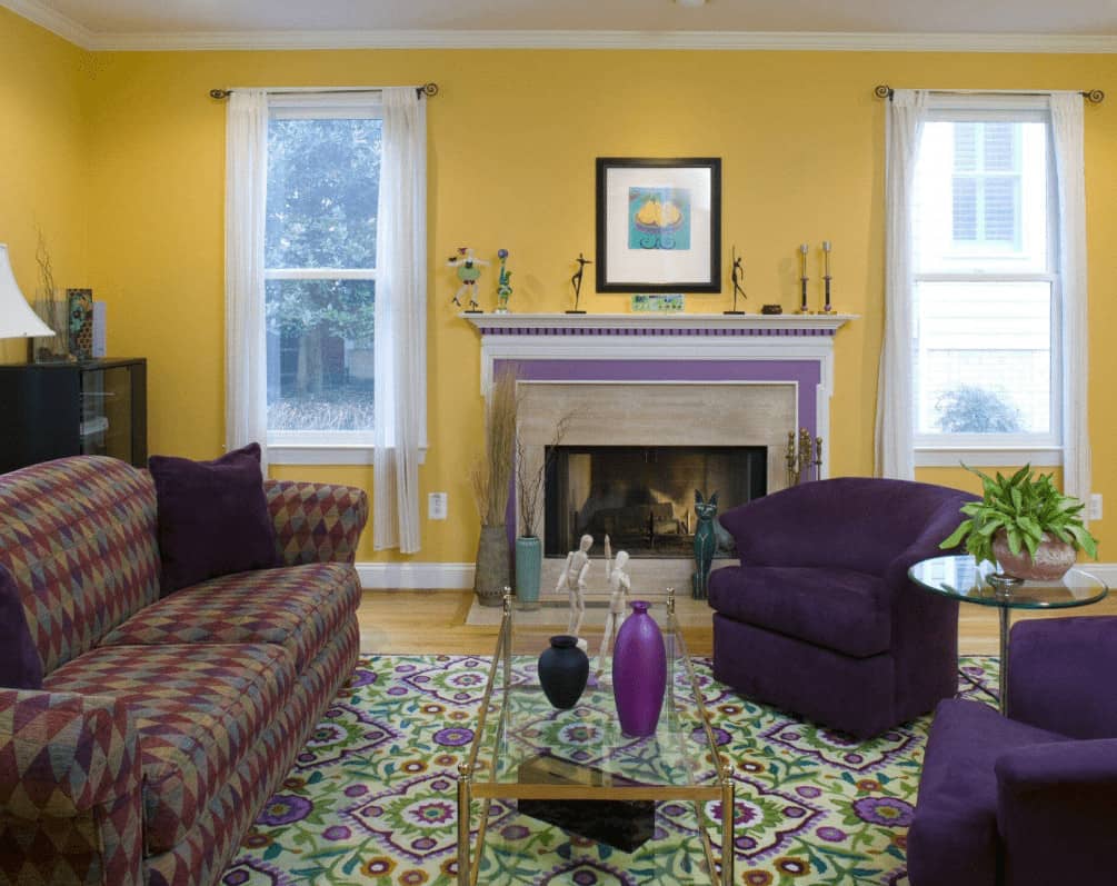 黄色的圆靠背椅与装有白色框架窗户的黄色墙壁形成了良好的对比。这间房间里有一张带图案的沙发和一张玻璃咖啡桌，还有一座壁炉，壁炉顶上摆放着装饰品和一幅黑色框架的艺术品。