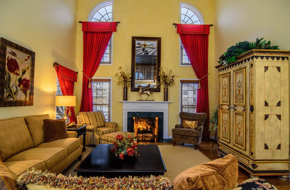 在这间黄色的客厅里，覆盖着玻璃窗的优雅红色窗帘格外显眼，客厅里有舒适的座椅和壁炉，壁炉上方摆放着花瓶和马雕塑。
