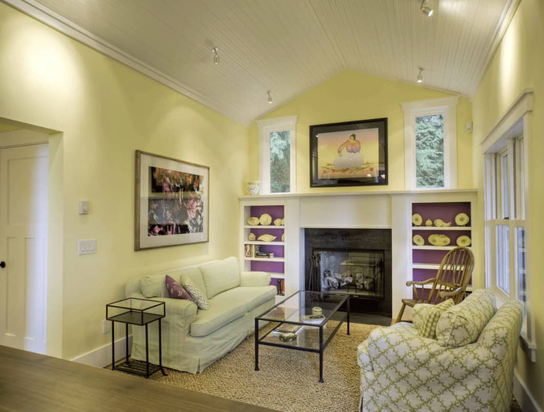 黄色的客厅，有舒适的座椅和一个玻璃封闭的壁炉，两侧是紫色的搁板。它由安装在教堂天花板上的镀铬轨道灯照明。