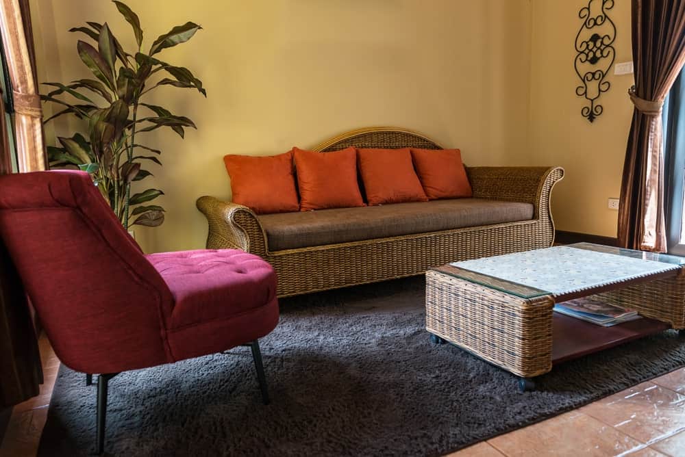 一把洋红色的簇绒椅子与华丽的墙壁艺术和柳条咖啡桌一起为客厅增添了华丽的色彩，这张咖啡桌与配有棕色靠垫和橙色枕头的沙发相得益彰。