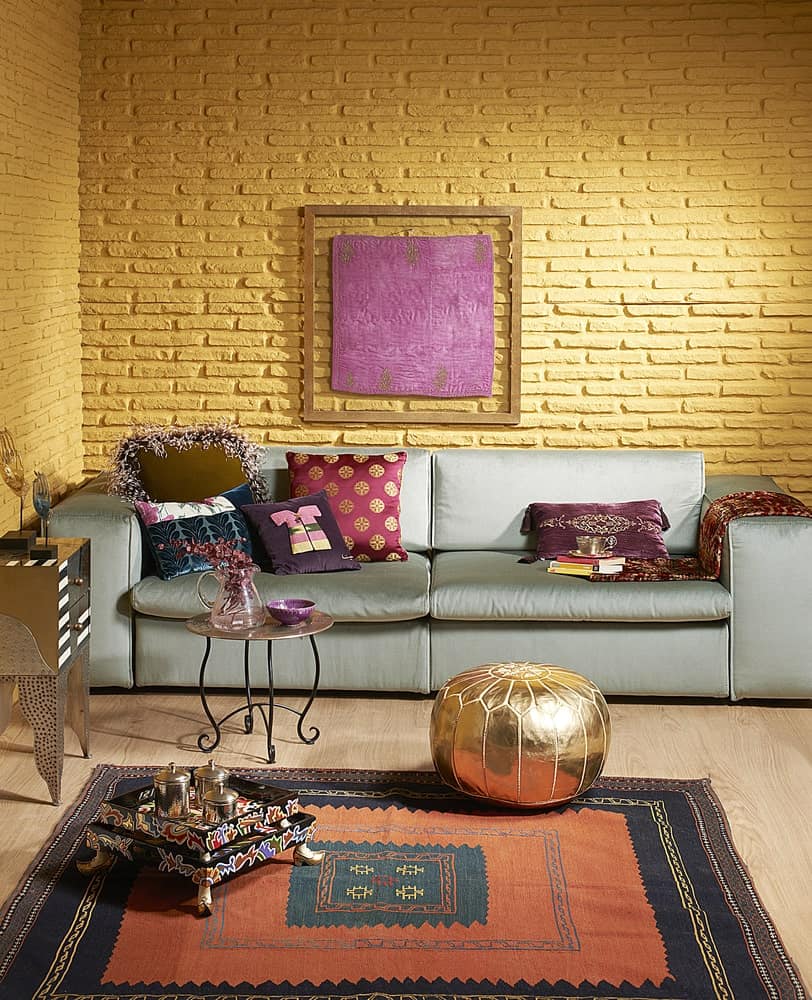黄色砖墙上挂着一幅木框墙画，上方是铺满华丽枕头的灰色沙发。它还配有一张金属咖啡桌和金色的软凳，搁在一块镶边的地毯上。