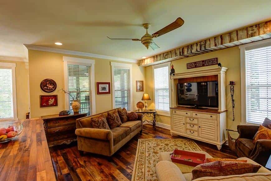 黄色的客厅，舒适的座椅和铺在硬木地板上的花卉地毯。它包括一个吊扇和放置在白色支架上的平板电视。