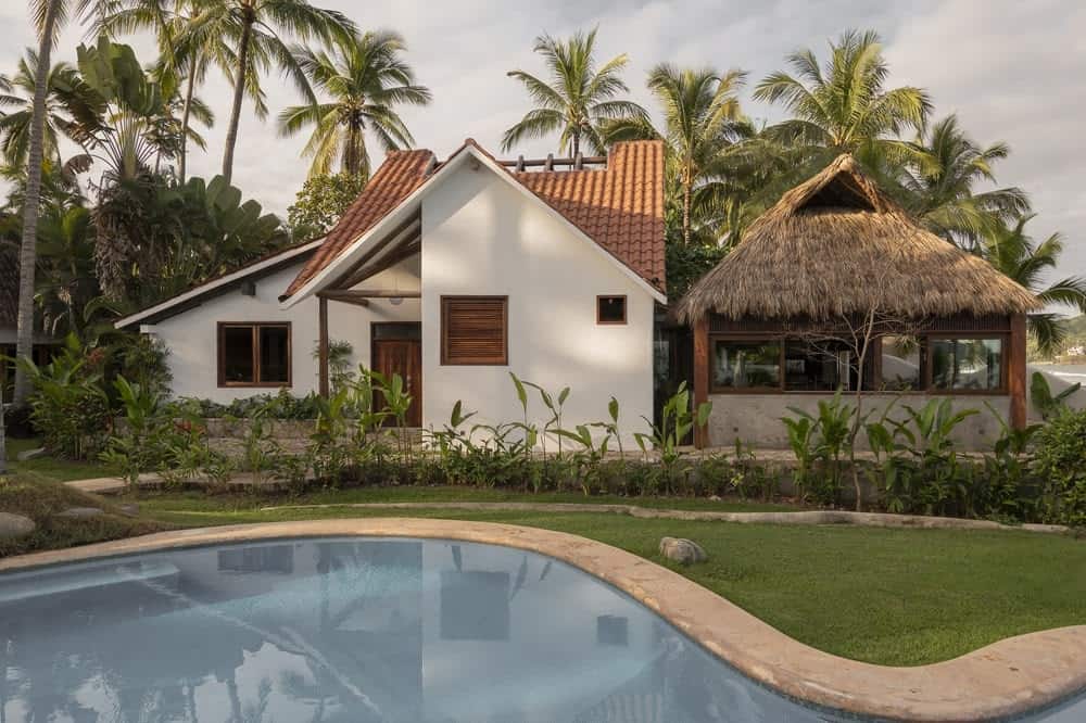 这是一座热带风格的住宅的全貌，它有明亮的墙壁，一面是红色的粘土瓦屋顶，另一侧是混凝土基础和竹框架的棕榈小屋。然后辅以周围高大的热带树木。