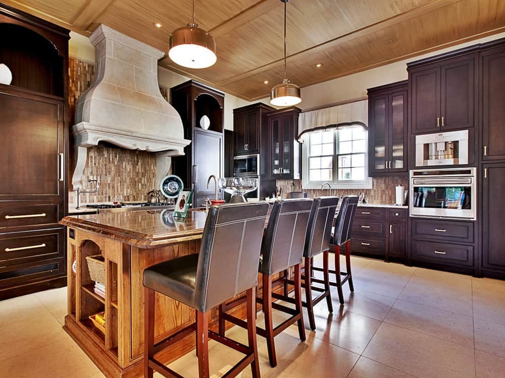 这个华丽的厨房有可爱的米色地板瓷砖，与木制厨房岛岛相辅相成，厨房岛岛配有棕色皮革凳子，顶部装有鼓形吊灯，来自露出横梁的木质天花板。