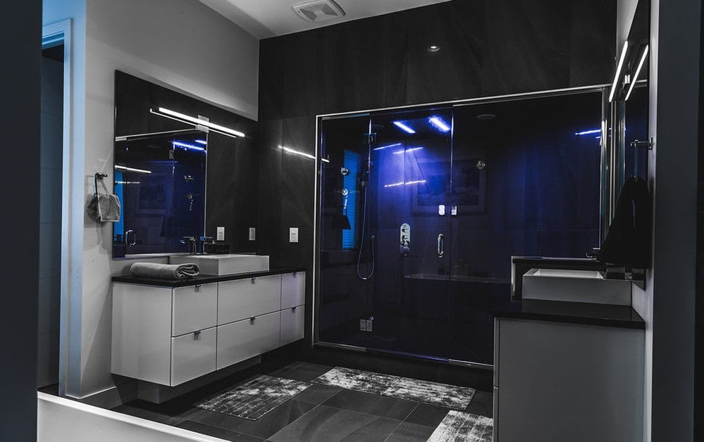 黑色的主浴室展示了一个步入式淋浴间，带有蓝色条形灯，以及由安装在无框镜子上的线性烛台照亮的面向容器的水槽梳妆台。