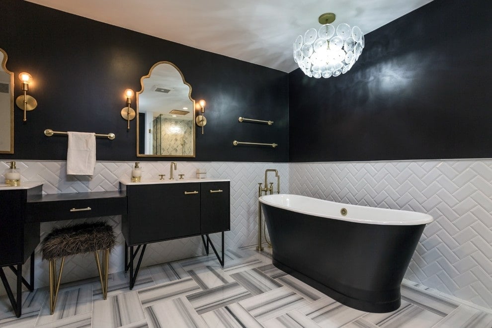 一个人造皮毛的凳子放在双洗手池梳妆台上，配有华丽的拱形镜子。旁边有一个黑色的独立浴缸，由黄铜枝形吊灯照明。