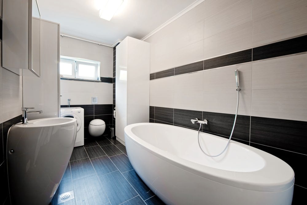 一个时尚的底座水槽正对着独立的浴缸，靠着黑白瓷砖的墙壁。它伴随着一个镜像药柜和一个前负载<a class=