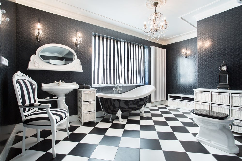 这个主浴室覆盖着黑色图案的壁纸，拥有一个基座式水槽和一个条纹软垫椅子，与爪足浴缸上方的窗帘相匹配。它包括一盏别致的枝形吊灯和开放的搁板，搁在布满储物箱的格子地板上。