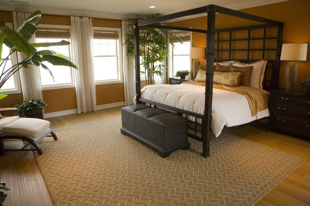 一间带有舒适床的主卧室，由台灯照明。房间里有室内植物和硬木地板，上面铺着一块小地毯。