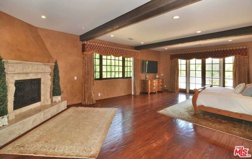 宽敞的主卧室，硬木地板和带横梁的天花板。房间里有一张漂亮的床和一座被棕色墙壁包围的壁炉。