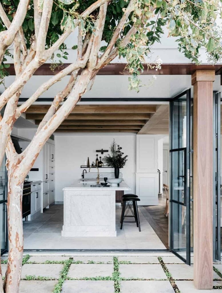 这是从敞开的玻璃门外看到的漂亮厨房。在这里，你可以看到明亮的白色色调主导着地板、墙壁和橱柜，与裸露的木梁相匹配的木制天花板相平衡。