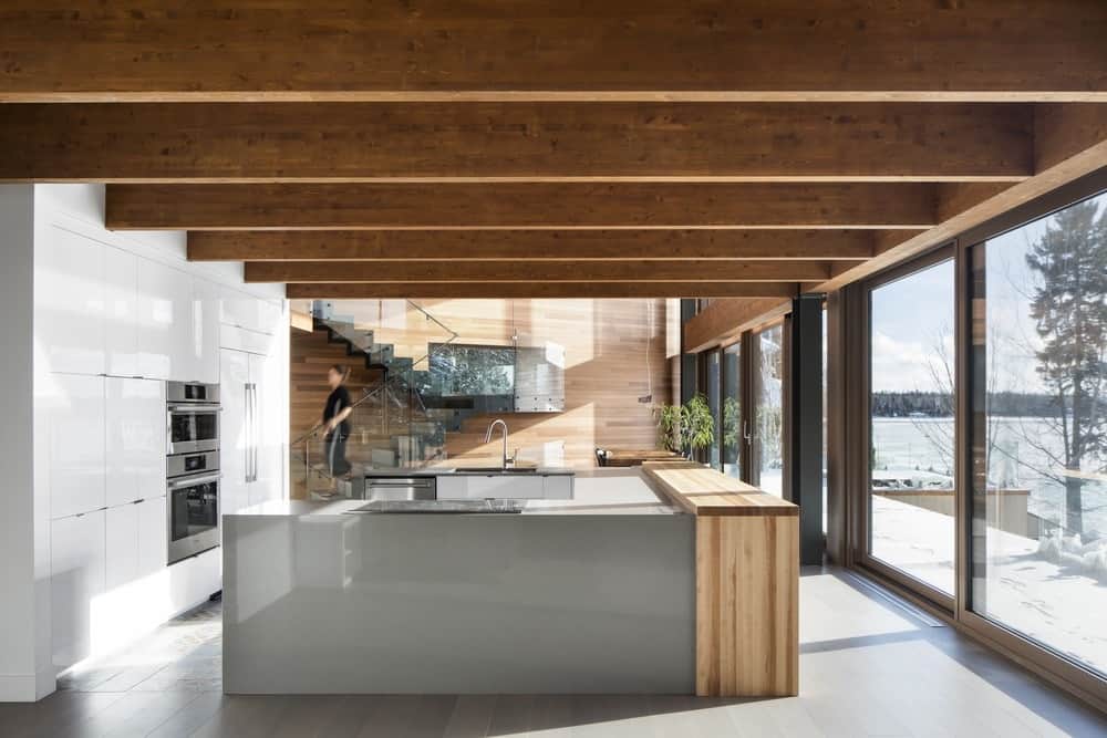 这是一个现代厨房，橱柜表面光滑光滑，中间有一个大的u形厨房岛。侧面的大型玻璃推拉门和露出木梁的宽木天花板增强了这些功能。