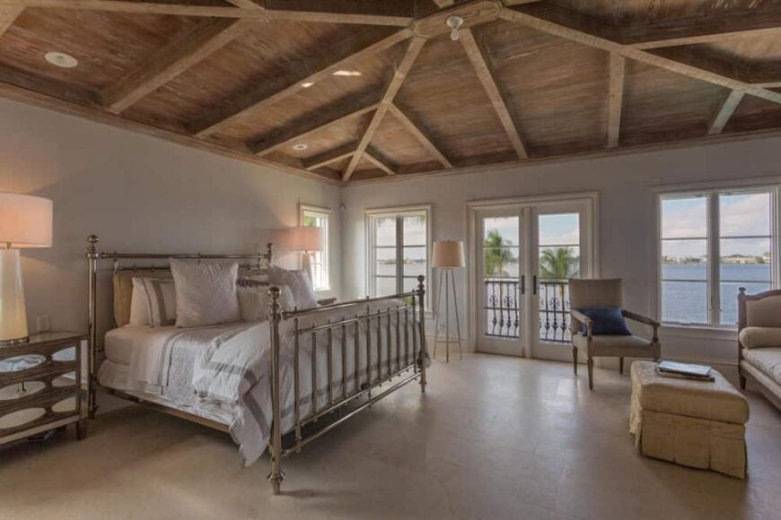 这是一个迷人而舒适的主卧室，四柱床上有金属栏杆，上面有舒适的白色靠垫和枕头。在房间的另一侧还有一个安静的休息区，它被统一的木质天花板和裸露的木梁抬高。