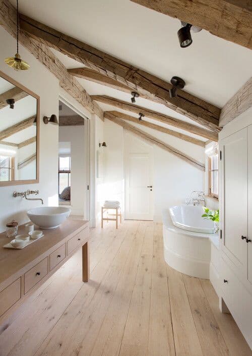 小屋天花板的质朴纹理木梁与浅色硬木地板相得益彰。这与木制的控制台桌子相匹配，作为碗水槽的虚荣。它的对面是连接浴缸的白色木柜。