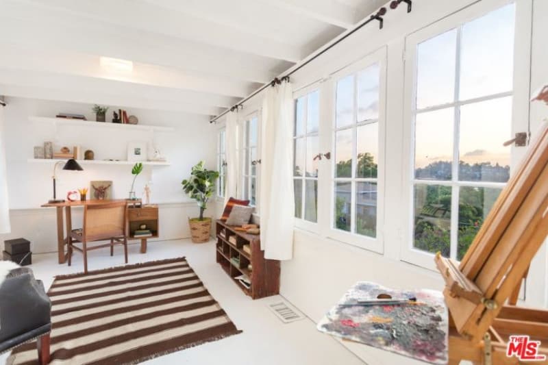 在这间白色的家庭办公室里，一条条纹地毯格外显眼，天花板上有横梁，窗框上覆盖着白色窗帘。它包括一张木桌和架子，上面有彩色的枕头。