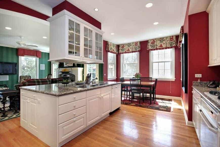 白色橱柜与厨房里的红墙形成了光滑的对比，厨房里有浅色硬木地板和白色框窗，窗户上装饰着印花帷幔。它包括花岗岩台面和一套木制餐具，放在经典的区域地毯上。