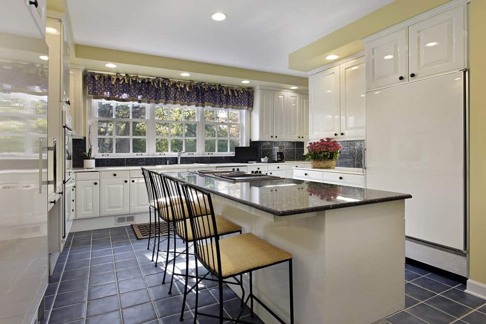 光滑的厨房配有黑色瓷砖地板和白色框窗，装饰着点缀的帷幔。它包括白色橱柜和配套的岛式酒吧，内衬有软垫金属椅子。