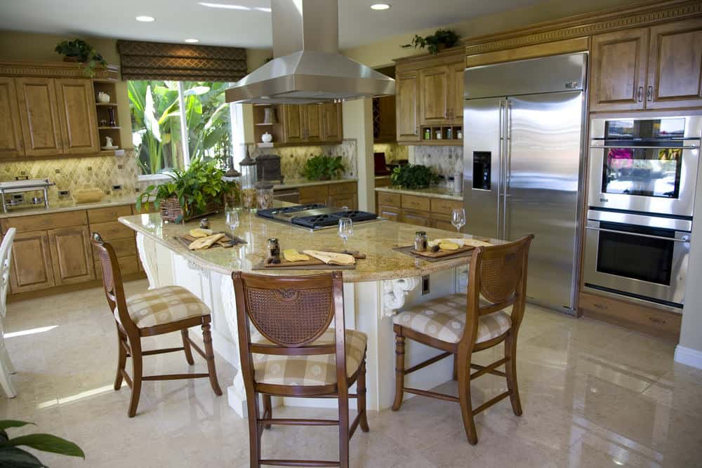 白色的厨房岛台配有木制软垫椅，上面有花岗岩台面和内置灶台。它被天然木质橱柜和不锈钢器具包围着。