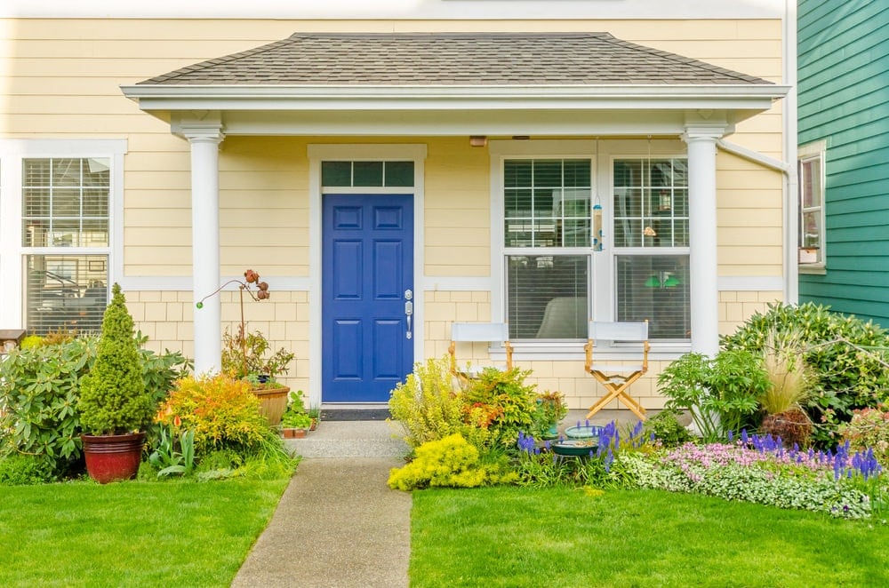 由郁郁葱葱的绿色草坪夹在一起的一条直接路面导致蓝色前门衬有白色柱的开放式门廊。它通过折叠浅黄色墙壁折叠椅子。