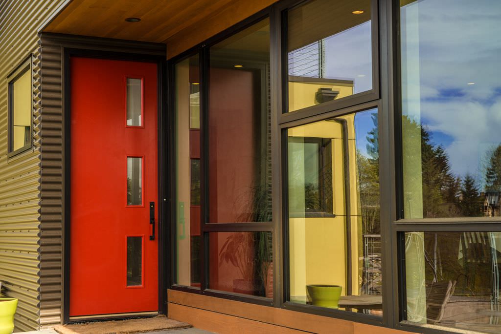 一扇光滑的红色门镶嵌着玻璃，铁艺把手在这座现代住宅中脱颖而出，展示了灰色的墙壁和玻璃镶板窗户。