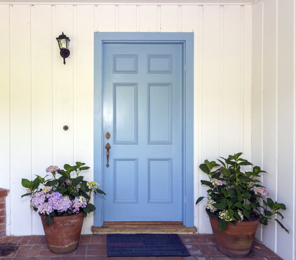 天蓝色的前门，黄铜把手，被安装在白色串珠板上的壁灯照亮。它还配有盆栽植物和一块深蓝色的地毯，上面铺着赤陶土地板。