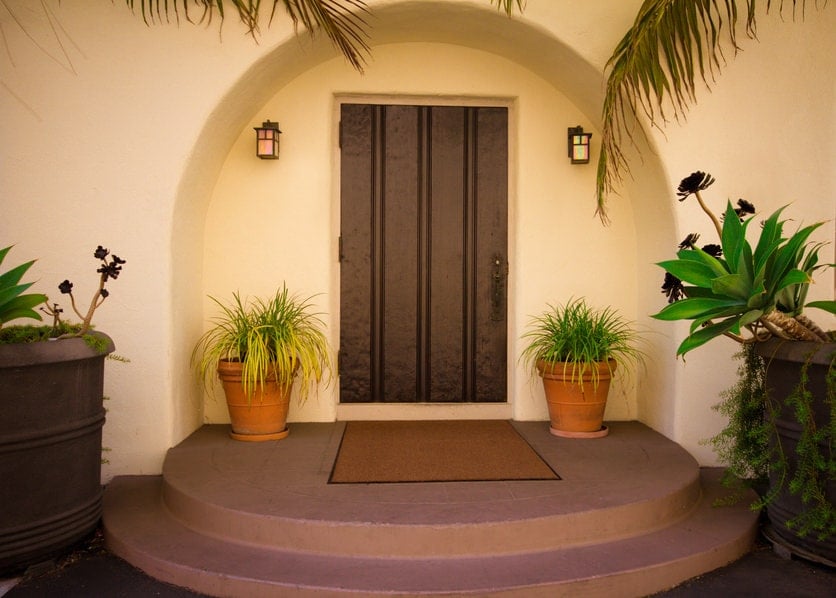一个简单的木制前门在一个拱形的嵌墙被户外的烛台照明。绿色的盆栽植物和混凝土平台上的棕色镶边地毯为它提供了补充。
