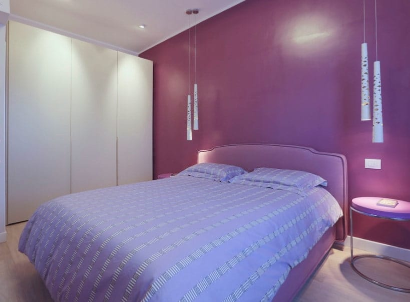 主卧室里舒适的大床被吊灯照亮。房间有一面紫色的墙和硬木地板。
