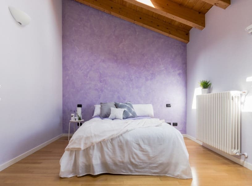 主卧室设有白色床，周围环绕着白色和紫色的墙壁，以及带有裸露横梁和天窗的木质天花板。