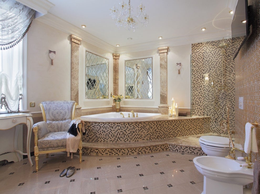 豪华的主浴室设计有时尚的镜子和一个花哨的水晶吊灯，挂在角落的浴缸覆盖着令人惊叹的马赛克瓷砖。它配有一张带图案的扶手椅和一个壁挂式电视下的卫生间。