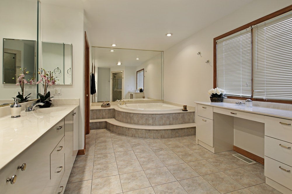 宽敞的主浴室，米色瓷砖地板和木制框架窗户覆盖白色卷帘。它包括面向镜子的梳妆台和一个角落里的浴缸。
