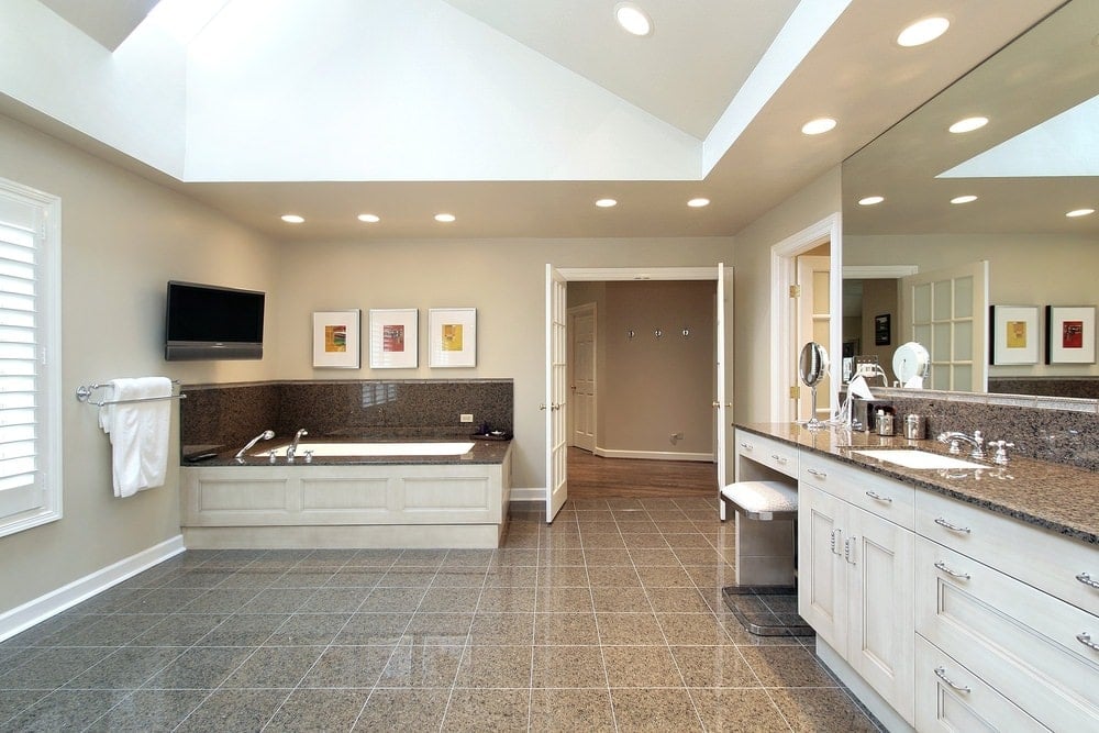 宽敞的主浴室，花岗岩地板和拱形天花板配有嵌入式灯。它包括一个大型洗手池梳妆台和一个深浴缸，浴缸固定在墙上安装的电视和白色框的艺术品。