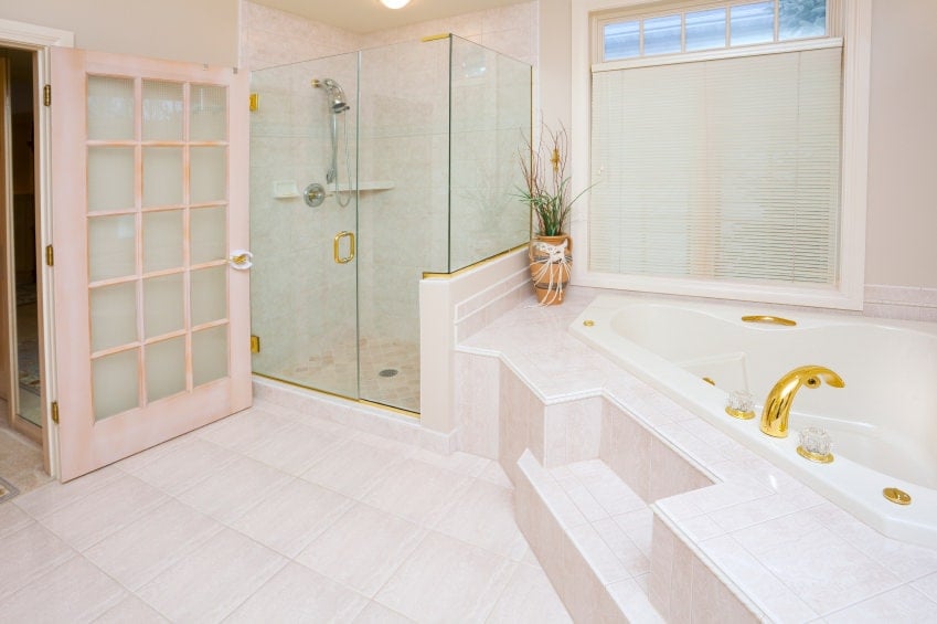 一扇玻璃门通往这间明亮的主浴室，其中有一间步入式淋浴间和一个角落里的金色浴缸。它有奶油色的地板和挂着白色卷帘的落地窗。