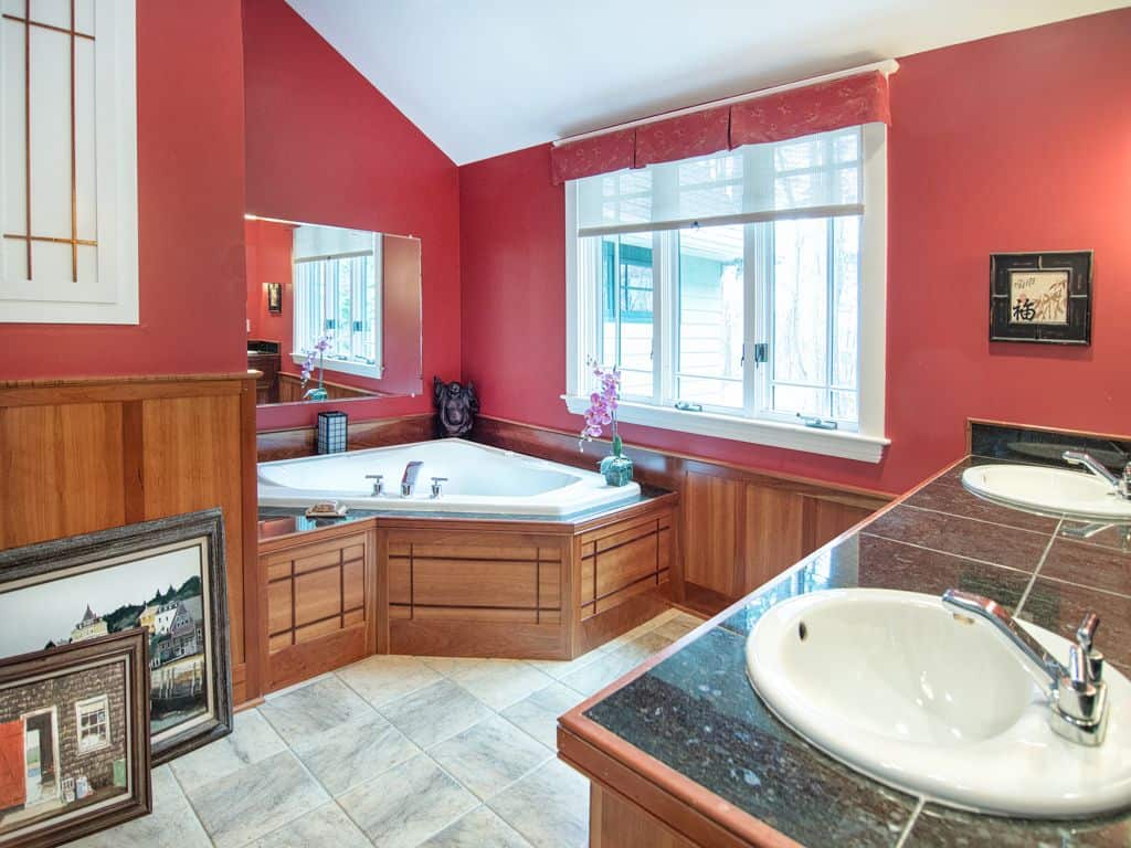 双水槽梳妆台面对着角落里的浴缸，浴缸覆盖着木板，与护壁板融为一体。这间主浴室装饰着华丽的艺术品，白色的窗框上挂着红色的帷幔。