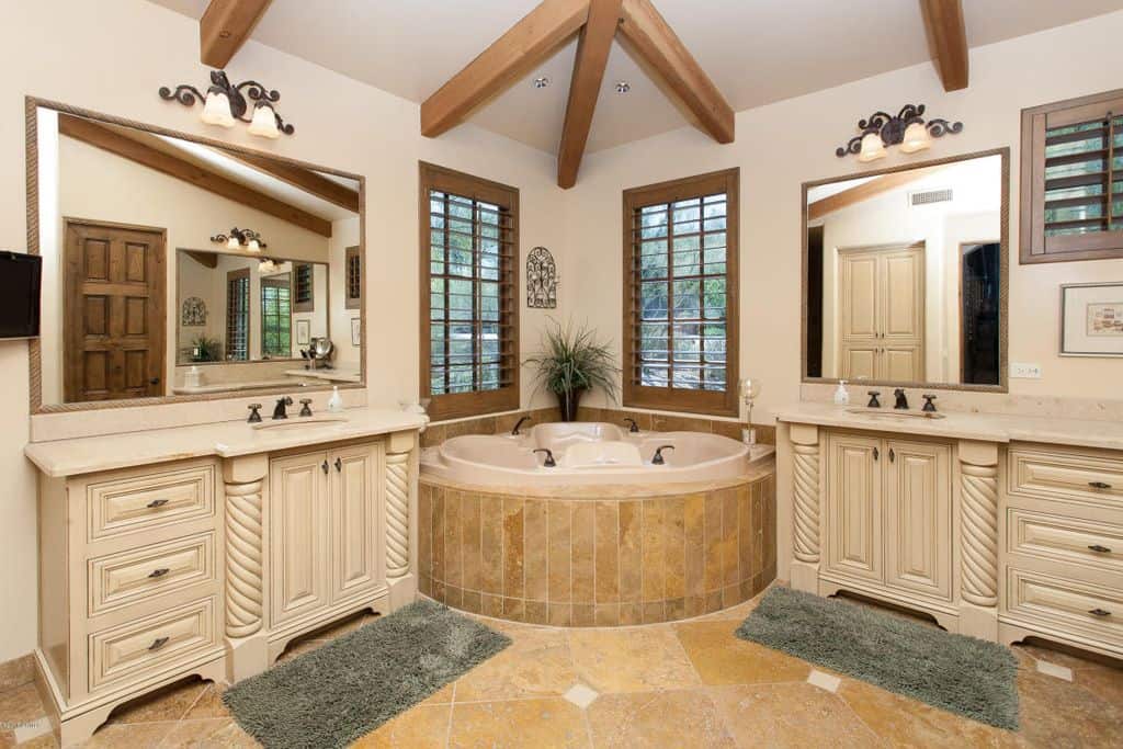 深浴缸是这个主要浴室的焦点，瓷砖地板和常规的白色天花板内衬天然木梁。它包括一对灰色的地毯，白色的梳妆台和木框窗户，在房间里创造出完美的对称。
