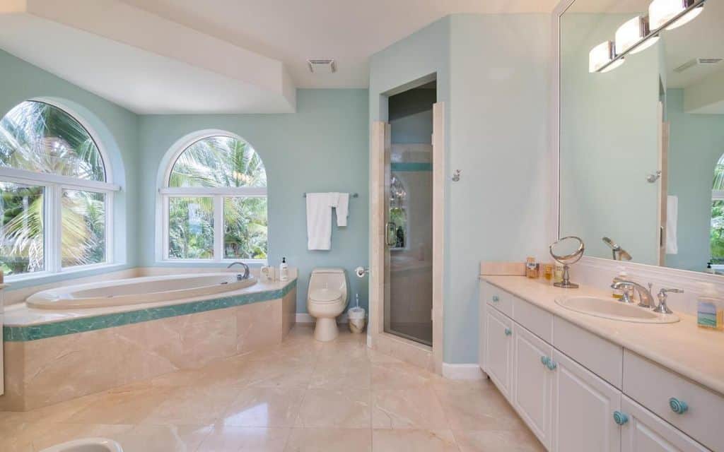 迷人的主浴室带有白色梳妆台，步入式淋浴间和拱形窗户旁的角落浴缸。它覆盖着米色大理石瓷砖，蓝色的色调与墙壁和把手相呼应。