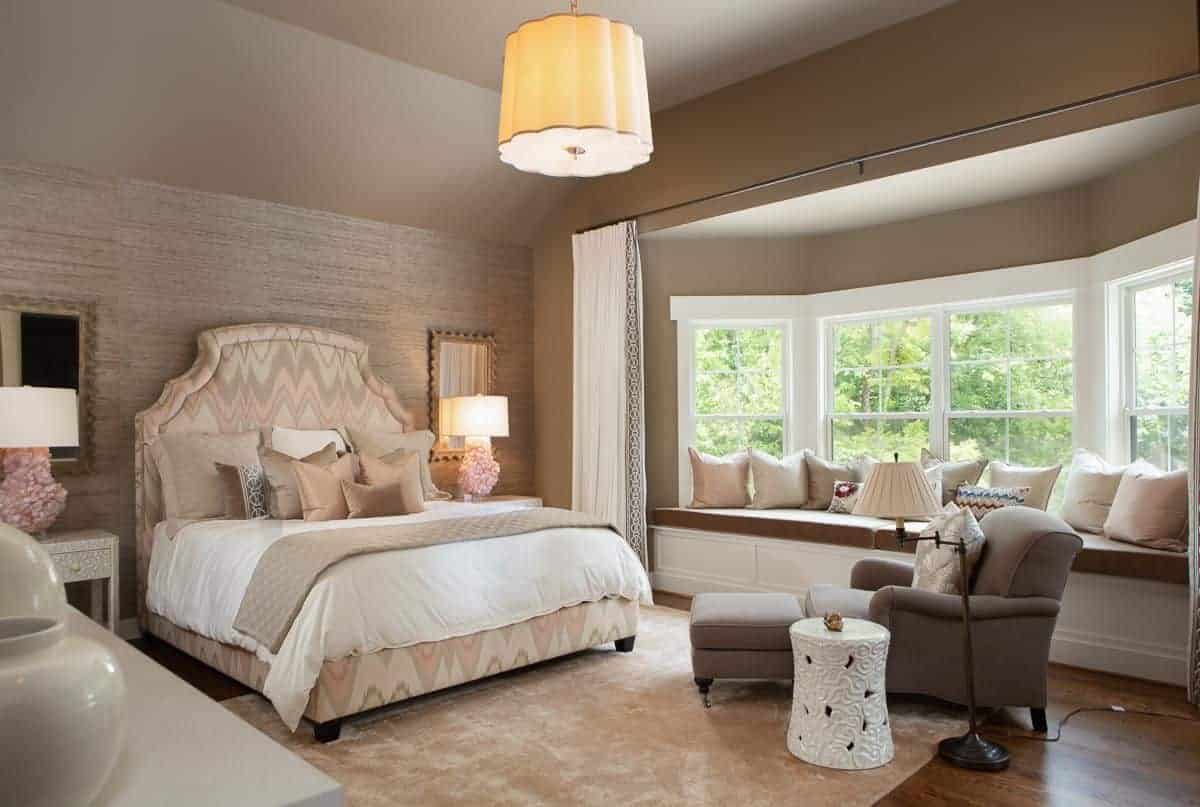 迷人的主卧室展示了一张可爱的图案床和一个靠窗的座位角落，配有棕色靠垫和中性枕头。它包括华丽的台灯和灰色的躺椅，搭配白色的边桌。