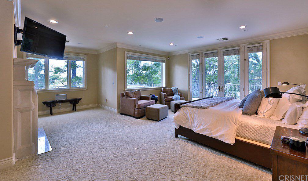 这间米黄色的主卧铺有纹理地毯地板，有一扇法式双开门通往阳台。它包括一台壁挂式电视和一张面向棕色扶手椅的深色木床，并配有配套的沙发。