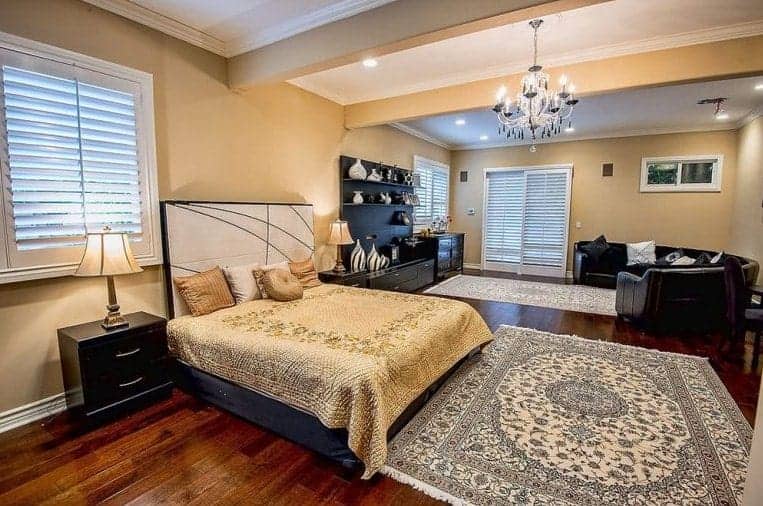 米黄色的主卧室里有嵌入式灯和一盏从横梁天花板上悬挂下来的华丽枝形吊灯。它有一张舒适的床和经典的地毯，还有一张弯曲的沙发，与黑色的床头柜和橱柜相得益彰。