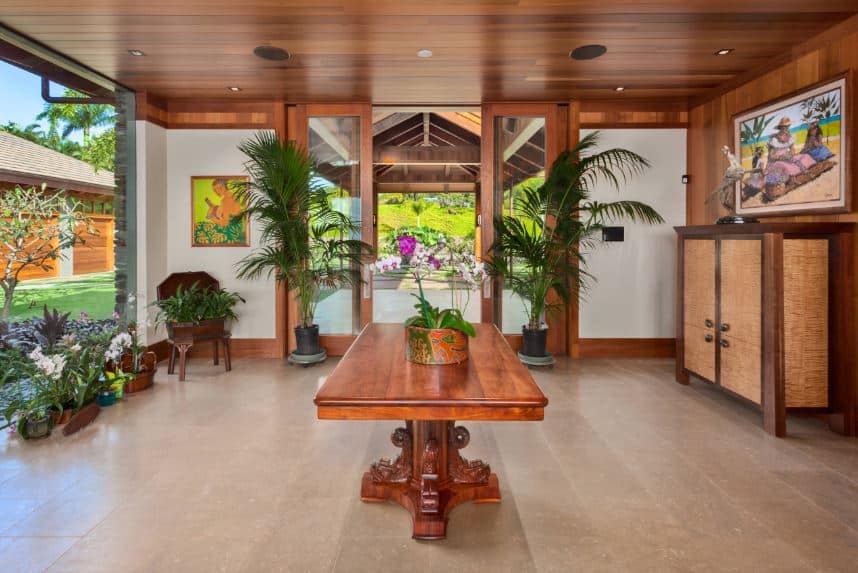 在这间热带风格的门厅里，米黄色地板中间的大红木桌子与天花板和门框的木质色调相匹配。门厅的亮度来自左边的大玻璃墙，与里面的各种盆栽热带植物很好地配合。